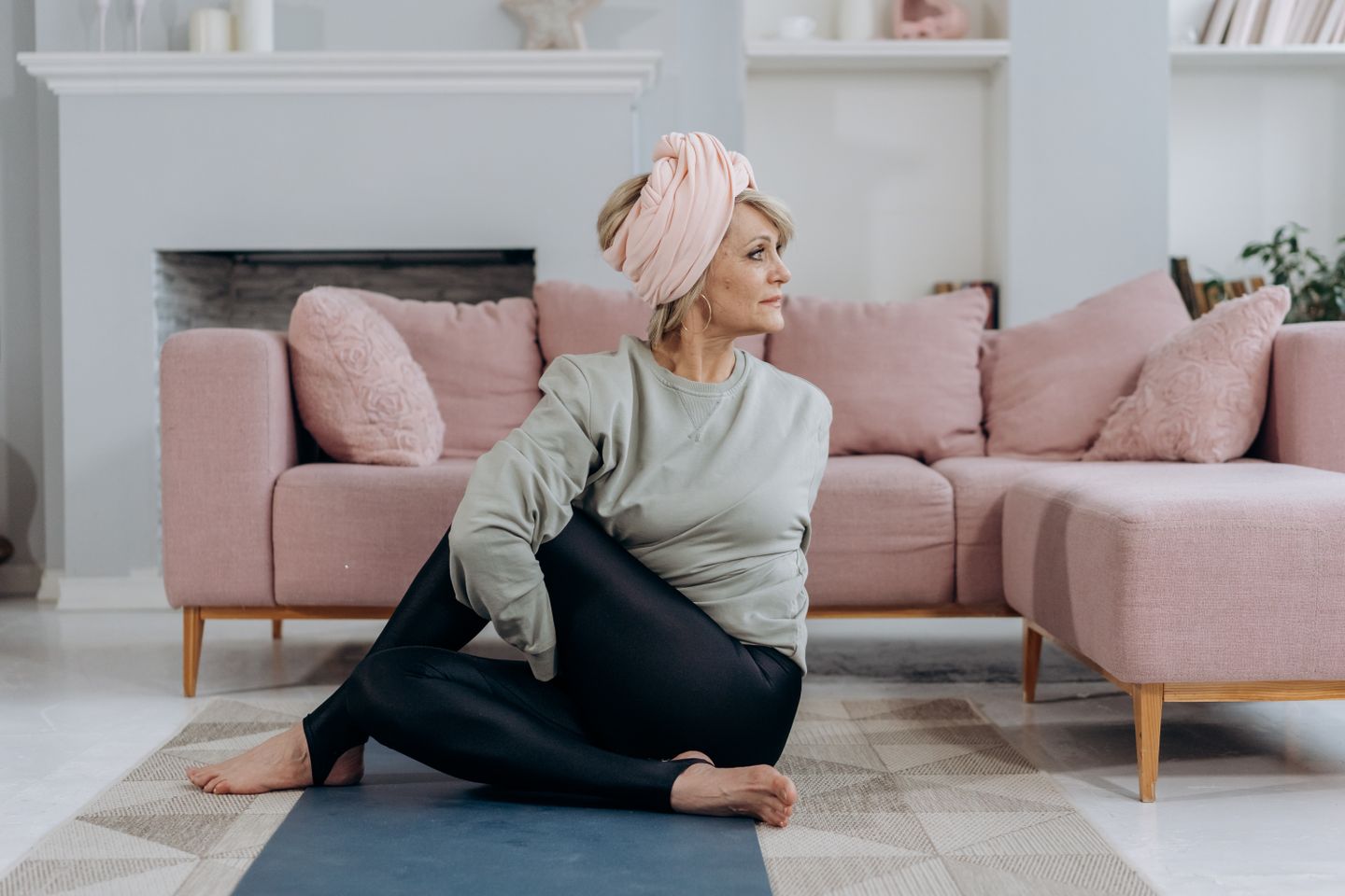 Full body of middle aged woman in sportswear and headband sitting on mat while stretching back near couch with pillows in light living room
